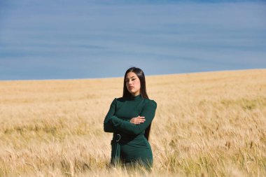 Yeşil giyinmiş, uzun siyah saçlı, güzel bir genç Latin kız bir yaz günü kollarını kuru sarı buğday tarlasında kavuşturur. Arka planda mavi bir gökyüzü var