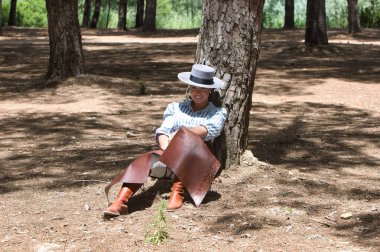 Genç, güzel bir kadın at kıyafetleri ve binicilik kıyafetleri giyer ve tipik bir İspanyol şapkası İspanya 'da kutsal bir günde bir çam ağacının gölgesinde dinlenir.