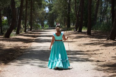 Geleneksel yeşil flamenko elbisesi giymiş, saçında bir çiçekle genç ve güzel bir İspanyol kadın kutsal yolculukta çam ağaçlarının arasında bir çubuğa yaslanarak yürüyor. Fotoğraf arkadan çekilmiş.