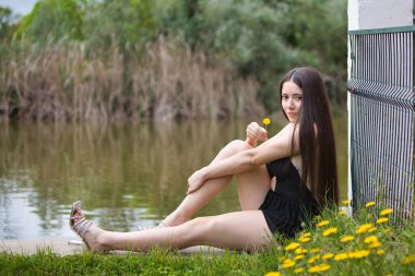 Siyah giyinmiş genç, esmer ve güzel bir kadın elinde sarı bir çiçek tutuyor ve göl kenarında yerde sarı çiçekler ve çimenler arasında mutlu bir şekilde oturuyor. Arka planda bir göl.