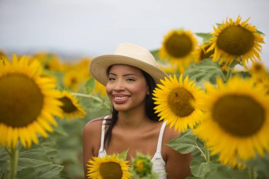 Beyaz bir bluz ve hasır şapka giyen güzel siyah bir Latin kadın ayçiçeklerinin arasında duruyor. Kadın mutlu ve gülümsüyor, kırsalda güneşli bir yaz gününün tadını çıkarıyor.