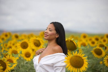 Koyu tenli, beyaz gömlekli ve çıplak omuzlu güzel bir Latin kadın güneşli bir günde ayçiçeklerinin arasında duruyor. Şehvetli kadın gülümser ve mutlu görünür.