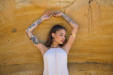 Makyaj yapan güzel bir genç kadın, bej bir elbise ve deniz kenarındaki bir uçurum duvarına dayanmış altın mücevherler. Kadın farklı pozlar ve ifadeler veriyor. Güzellik ve moda kavramı