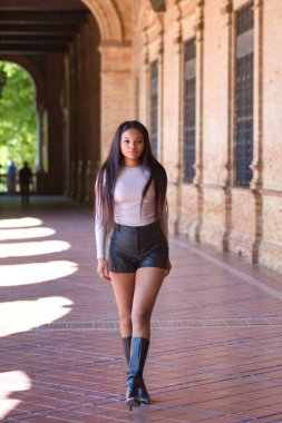 Deri şort, siyah çizme ve bej bluz giyen genç ve güzel Afrikalı kadın tatilde ve İspanya 'nın ünlü meydanında Seville' de yürüyor. Seyahat ve tatil konsepti