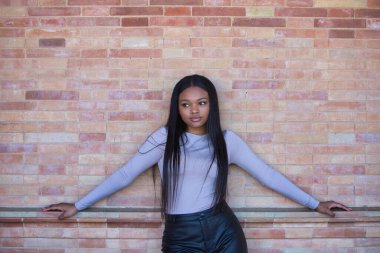 Afrikalı bir kadın, genç ve güzel, bej bluzlu ve siyah deri şortlu. Kolları açık, kırmızı tuğladan bir duvara yaslanmış.