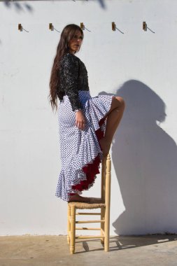 Latin kadın, genç ve güzel, dans eden flamenko eski bir tahta sandalyede kameraya bakıyor. Kadın, siyah benekli, kısa siyah ceketli, beyaz, dalgalı bir elbise giyiyor.