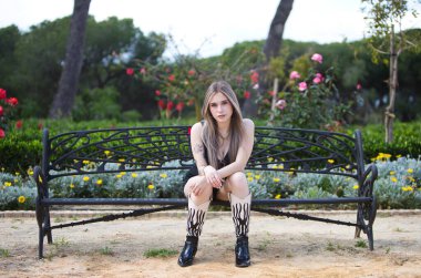 Genç, güzel, sarışın, dövmeli, siyah çizmeli, parktaki bankta oturan ve kolları bacaklarında dinlenen bir kadın.