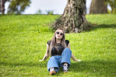 Genç, güzel, güneş gözlüklü, kot pantolonlu ve siyah gömlekli bir kadın çimlerin üzerinde bir ağacın gölgesinde oturuyor. Kadın ellerini dinlenmek için arkaya yaslıyor.