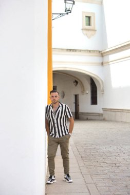 Genç, sakallı, çizgili gömlek ve kargo pantolonu giyen yakışıklı bir adam vücudunu Seville, Endülüs, İspanya 'nın ortasında kemerli beyaz bir duvara yaslıyor.
