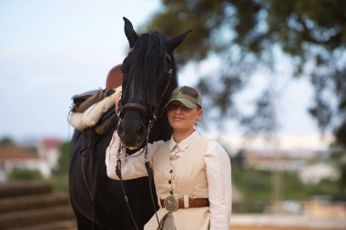 Genç ve güzel bir kadın siyah safkan İspanyol atının yüzünü sevgiyle tutuyor ve kameraya mutlulukla bakıyor. Kadın tipik bir atlı kadın kostümü giymiş.