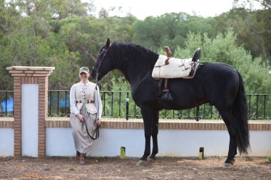 İspanya, Endülüs 'te siyah safkan İspanyol atının yanında bir çam ağacının gölgesinde oturan güzel genç kadın. Kadın tipik bir atlı kadın kostümü giymiş.
