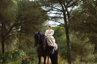 Genç ve güzel bir kadın çam ormanında safkan bir İspanyol siyah atına biniyor. Kadın tipik bir atlı kadın kostümü giyiyor. Arka planda yemyeşil bitki örtüsü