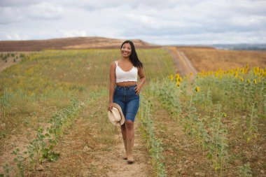 Beyaz bluzlu, kot pantolonlu ve hasır şapkalı güzel bir Latin çiftçi ayçiçeği tarlasını gözetliyor. Kadın bu güneşli yaz gününde mutlu ve gülümsüyor.