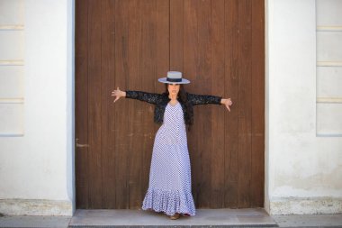 Latin bir kadın, Endülüs 'ün tipik bir sokağındaki ahşap bir kapının önünde dans eden genç ve güzel bir flamenko. Kadın, siyah benekli, şapkalı ve ceketli beyaz bir elbise giyiyor.