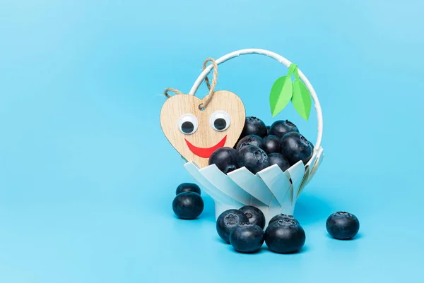 Yeşil yapraklı ev yapımı beyaz bir sepetin içinde taze çiğ yaban mersini ve mavi arka planda gülen tahta bir kalp..