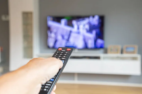 Arka planda bulanık bir televizyonu olan siyah bir TV kumandasını tutan bir erkeğe yakın çekim.. 