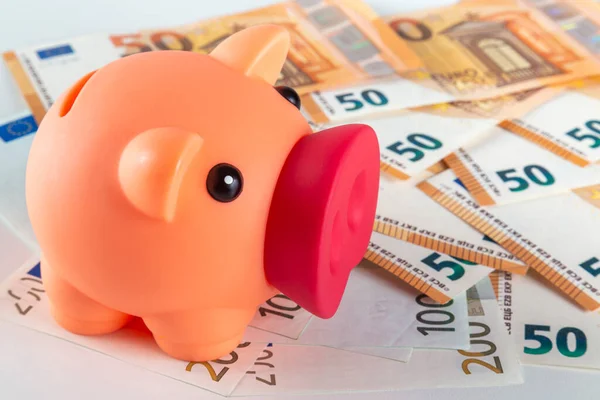A pink pig piggy bank stands on a table covered with large quantity of euro banknotes on a light background close-up