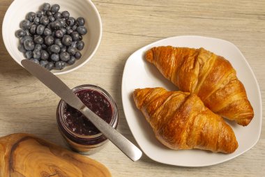 Beyaz tabakta iki altın kruvasan, bir kavanoz koyu meyve reçeli ve bir kase yaban mersini..