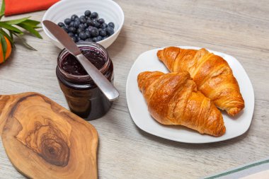 Beyaz bir tabakta iki kruvasan, bir kavanoz koyu meyve, bir bıçak ve bir kase yaban mersini..