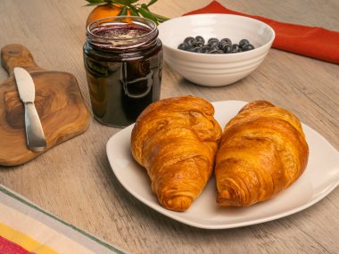 Kruvasan, reçel kavanozu, tereyağı bıçağı, ahşap kesim tahtası ve ahşap masada taze yabanmersini..