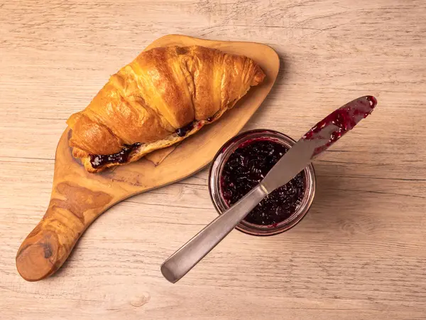 Tahtayı dolduran kruvasan ve tahta yüzeyde bıçaklı reçel kavanozu.