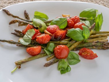 Beyaz tabakta taze fesleğen ve kiraz domatesli ızgara kuşkonmaz, sağlıklı ve renkli bir vejetaryen yemeği..