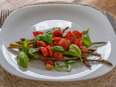 Çilekli domates ve fesleğen yapraklarıyla kaplı kavrulmuş kuşkonmaz, taze ve sağlıklı bir tabakta servis edilir..