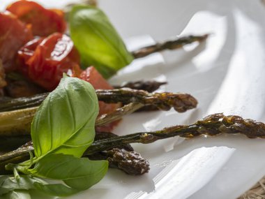 Taze ızgara kuşkonmaz, kavrulmuş kiraz domatesleri ve mis kokulu fesleğenle birlikte beyaz bir tabakta güzelce sunuluyor..
