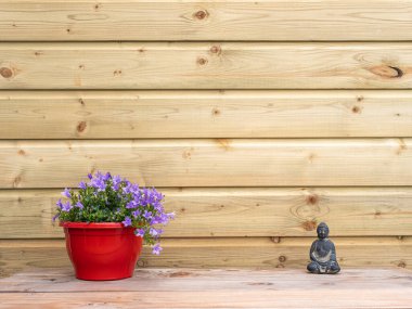 Kırmızı çömlekte mor çiçekler ve hafif ahşap duvara karşı bir Buda heykeli, asgari kırsal dekor..