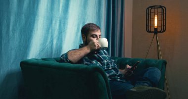 Man Drinking Coffee on a Green Soft Armchair. Relaxed Male Drinking Coffee. Coffee Drinking. Home. Green Sofa