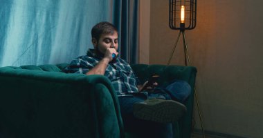 man vaping on a green armchair , man checking social media via phone. man inhaling an e-cigarette vaping device. smoking electronic cigarette vapor. Smoking Electronic Cigarette, Vape, Man Vaping