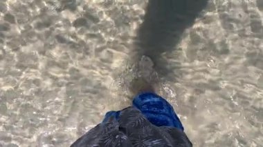 Çıplak ayaklı ıslak sahil kumu. Erkek bacakları ve ayakları kumlu sahilde deniz dalgaları boyunca yürüyor. Adam deniz kıyısında sörf yapıyor. Su ve köpük damlaları. yavaş çekim.