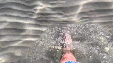 Çıplak ayaklı ıslak sahil kumu. Erkek bacakları ve ayakları kumlu sahilde deniz dalgaları boyunca yürüyor. Adam deniz kıyısında sörf yapıyor. Su ve köpük damlaları. yavaş çekim.