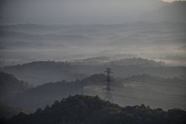 Sis bir vadiden akar ve dağın tepesinde elektrik iletim kuleleri vardır..