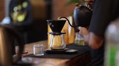 Barista kahve zeminine sıcak su dökerek kahve yapıyor. Bakır bir tencereden şeffaf bir krom damlatıcısına kadar hazırlanmış filtre ile kafeteryadaki ahşap bir masanın üzerine dökülüyor.