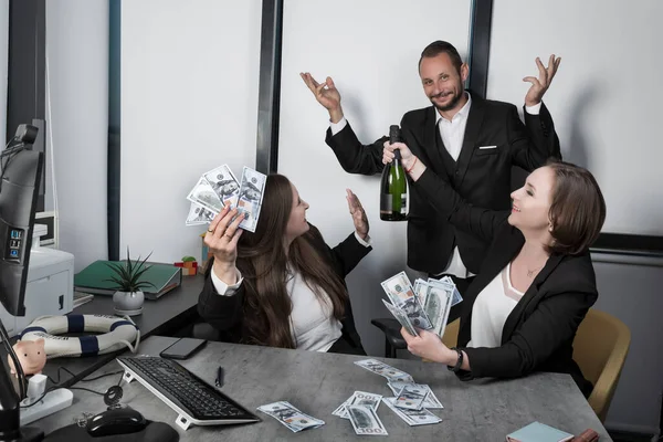 两个快乐的女人坐在桌旁 手里拿着一捆钱和酒瓶庆祝这一盛事 快乐兴奋的同事一起庆祝胜利在办公室与香槟 令人惊讶的人 — 图库照片