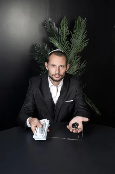Vertical photo of caucasian Jew israeli office worker holding a stack of dollars in hands. Bearded Jewish man in Yarmulke (hat, Kippah) sit at desk, show pocket watch in hand and hundred dollar bills.
