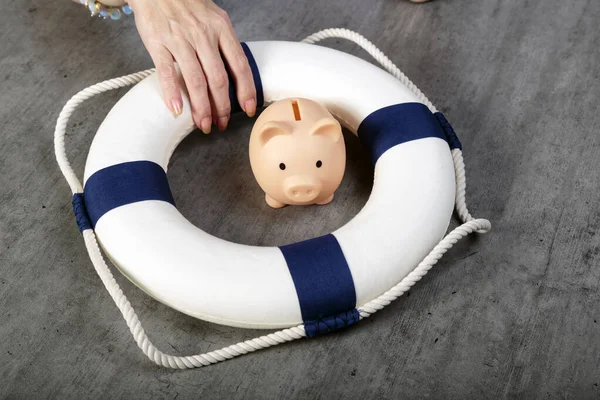 Woman hand with little piggy bank inside of lifebuoy. Safe Money. Blue and white life buoy on wooden desk table background. Assets wealth, money saving and security by insurance concept. Copy space.