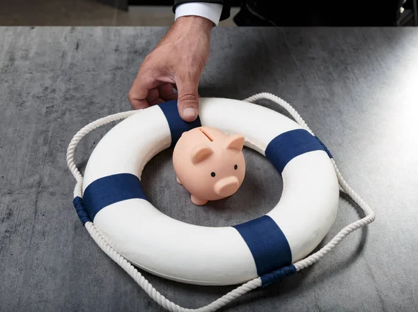 Male hand with little piggy bank inside of lifebuoy. Safe Money. Blue and white life buoy on wooden desk table background. Assets wealth, money saving and security by insurance concept. Copy space.