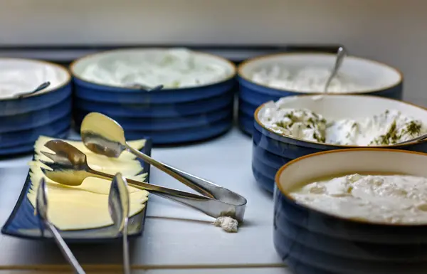 Cheese plate served in a blue plates with steel spoon tongs on table. Tasty cream cheese, bowl party platters. Slices of cheese and serving kitchen tongs on a bowls mayonnaise and yogurt background