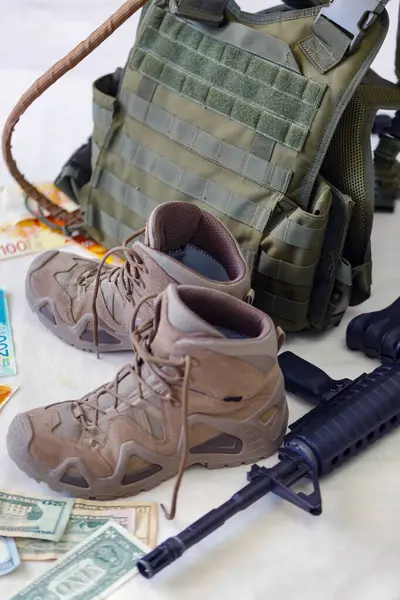 Bulletproof vest, money banknotes USA american dollar bills with israeli NIS , rifle and worn hiking boots on white floor. Military uniform protected with body armor, assault rifle, machine gun 16mm