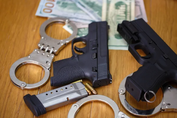 two semi automatic pistols with handcuffs, bullet shells 9 mm and American dollar banknotes on blurred background. Pistol gun, two police handcuffs and us dollars banknotes money on wooden table