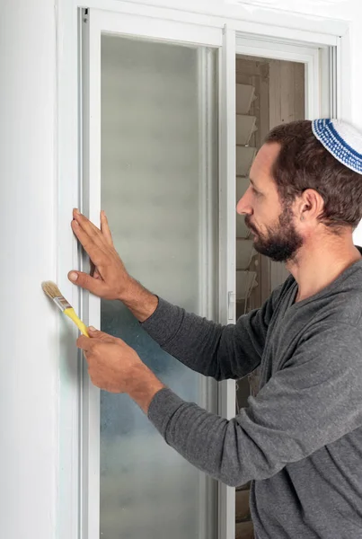 Handyman in skull cap painting wall near windows with a brush, repair. Painter painting the bottom of a window frame and an angled sash brush with close up of hand. Jew man in a kippa hat on his head