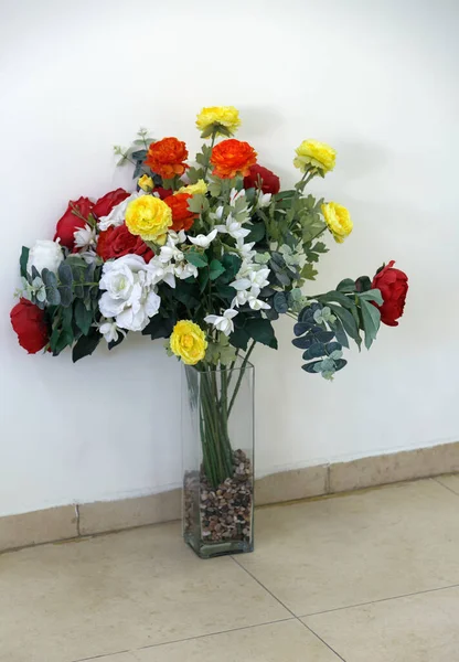 A transparent vase with flowers on floor on white wall background. Set of a high glass flowerpot and artificial bouquet of roses flowers in transparent bottle used to decorate in various ceremonies