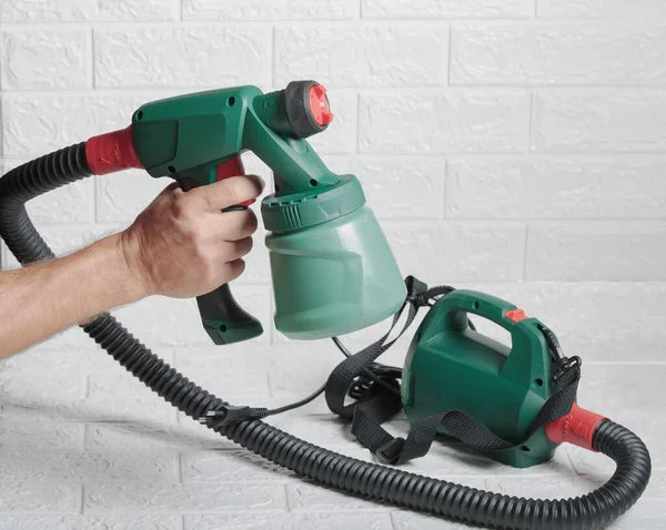 Hand Holding a Green Paint Sprayer Against a White Brick Wall. A person using a paint sprayer for home renovation. Electric colored paint sprayer, useful instrument for quick paint job. Spray gun