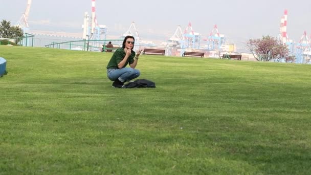 一个穿着休闲装的女人坐在公园里一片生机勃勃的绿地上 一个可爱的女孩坐在一个阳光灿烂的公园里 涂着口红 穿着绿色毛衣 牛仔裤和太阳镜 她的黑头发闪闪发亮 脸蛋漂亮 — 图库视频影像