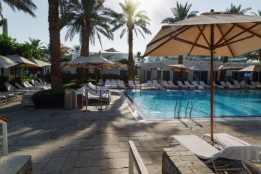 Parlak güneş ışığı, sıcak ve davetkar bir atmosfer yaratıyor. Beyaz şezlonglar havuzun etrafına güzelce yerleştirilmiş. Bazı sandalyelerin gölgesi bej şemsiyelerle kaplanarak güneşten korunuyor. Bazı sandalyeler bej renkli şemsiyelerle gölgeleniyor.