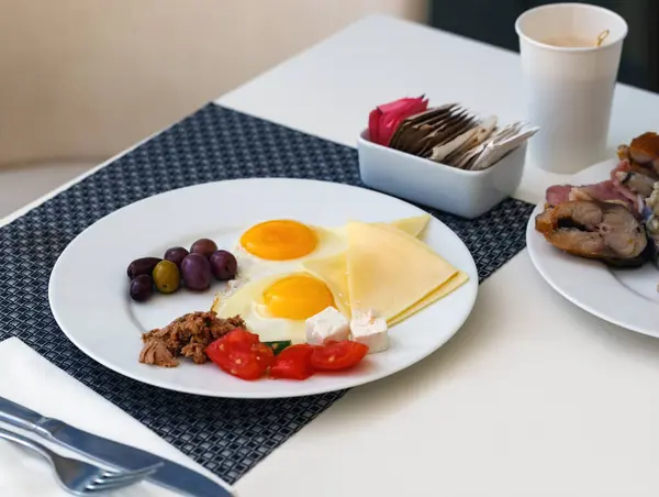 stock image A breakfast setting on a table covered with a dark textured tablecloth. A white round plate holds sunny side up eggs, cheese slices, olives, cherry tomatoes and mackerel fish, paper cup of hot coffee