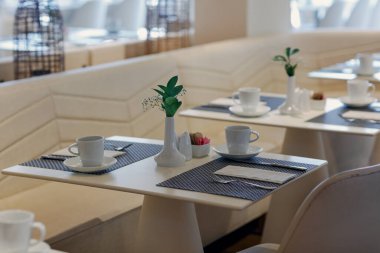 İki kişilik masaları olan yemek salonu. Her masa yeşil yapraklar ve nefes çiçekleri, fincanlar ve çay tabakları ve şeker tutacağı içeren beyaz bir vazoyla süslenmiştir..