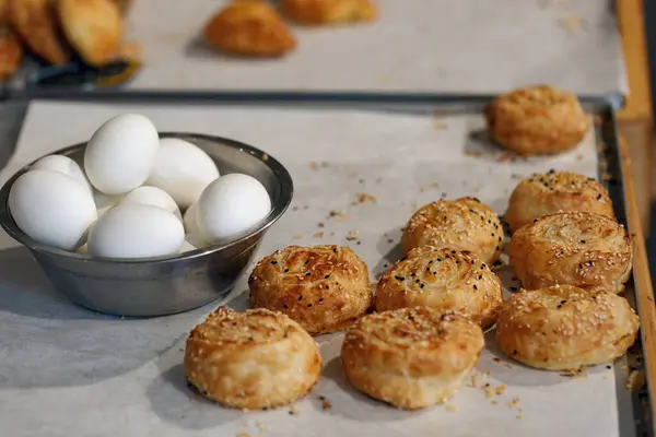 A baking sheet with golden brown rolls sprinkled with seeds. Next to the rolls, there\'s a bowl of white eggs. Israeli breakfast including baked pastry bourekas and egg. The scene suggests the preparation or aftermath of a baking session